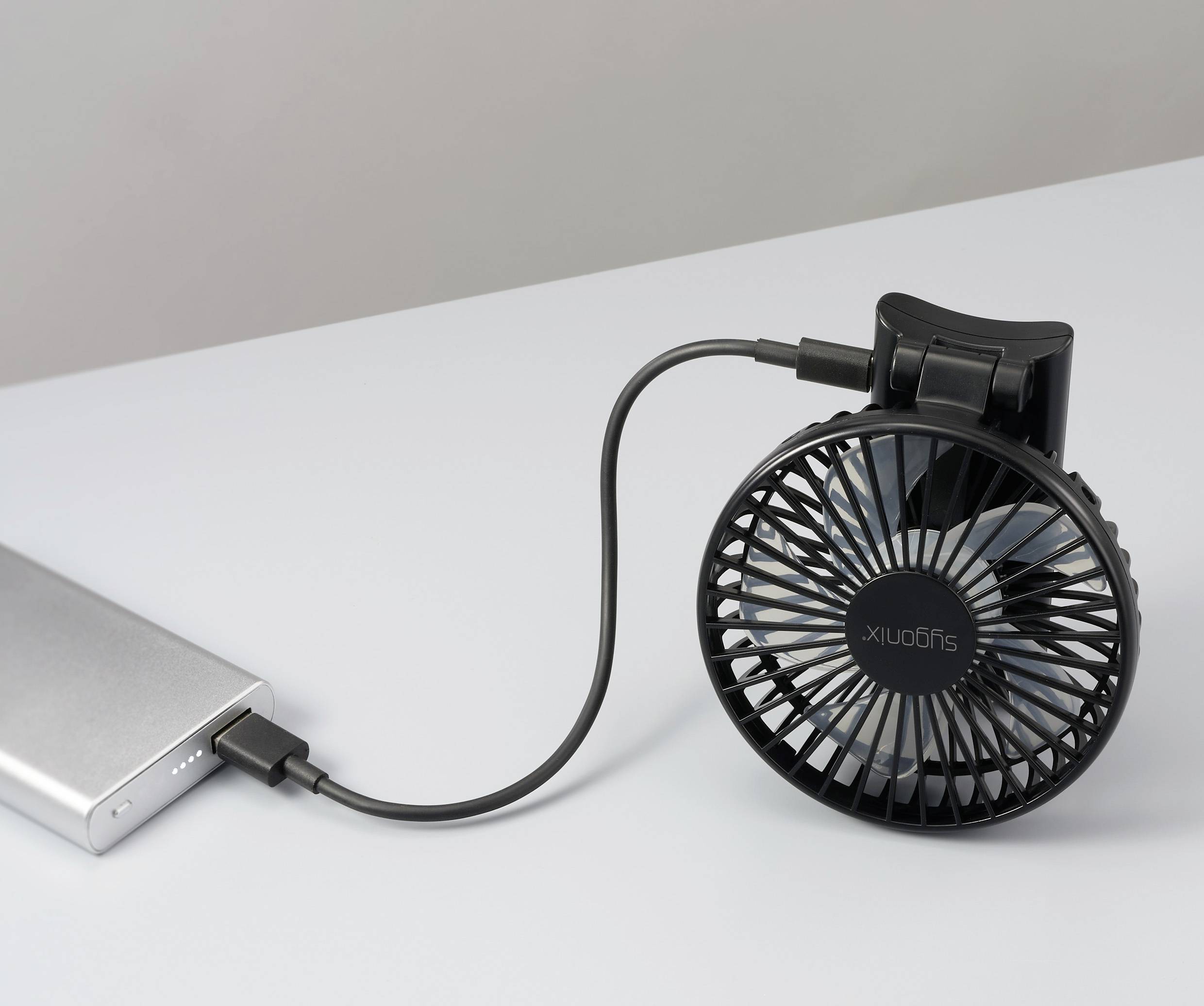 Un petit ventilateur noir relié à une batterie externe via un câble USB sur une surface blanche, démontrant sa portabilité et sa facilité d'utilisation.