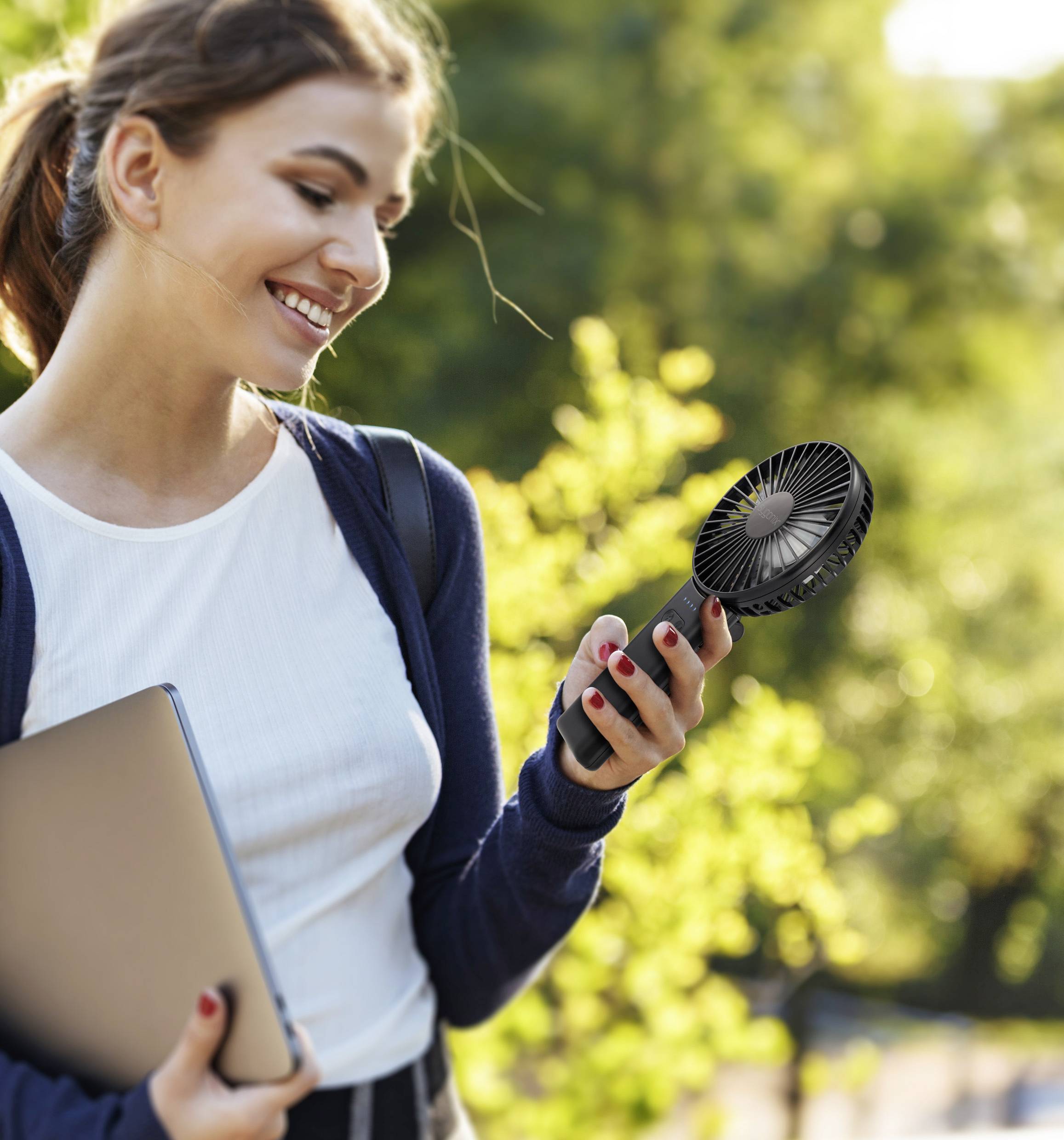 Une personne sourit tout en tenant un ventilateur portable et en transportant un ordinateur portable à l'extérieur dans un cadre verdoyant et ensoleillé.