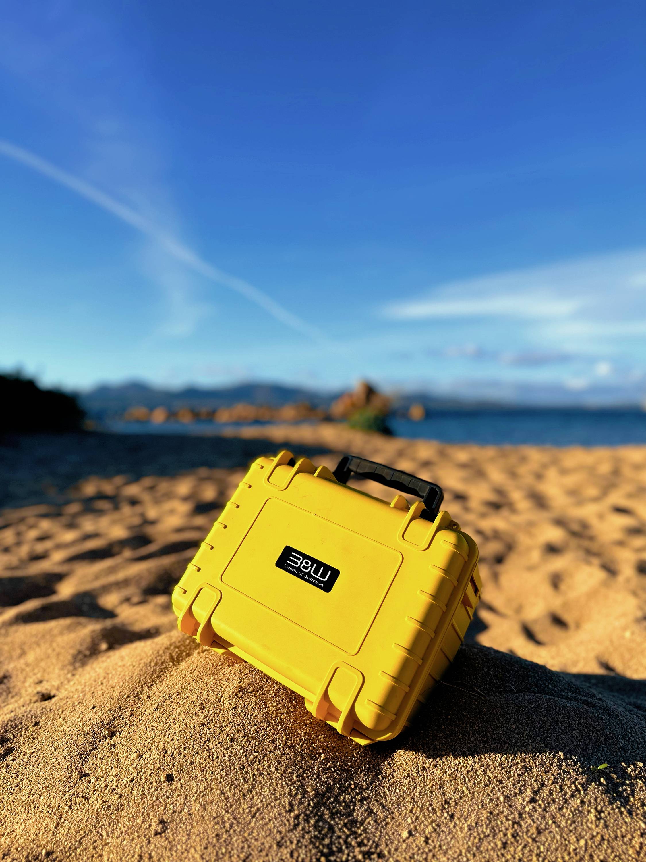 Un étui de protection jaune vif repose sur une plage sablonneuse sous un ciel bleu dégagé, avec des montagnes lointaines et la mer visibles en arrière-plan.