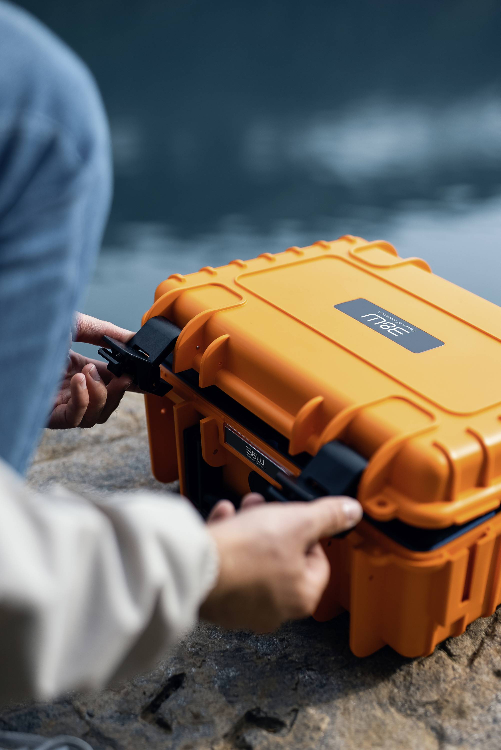 Une personne ouvre un étui en orange robuste et durable sur une surface rocheuse près de l'eau, suggérant une utilisation en extérieur ou lors de voyages.