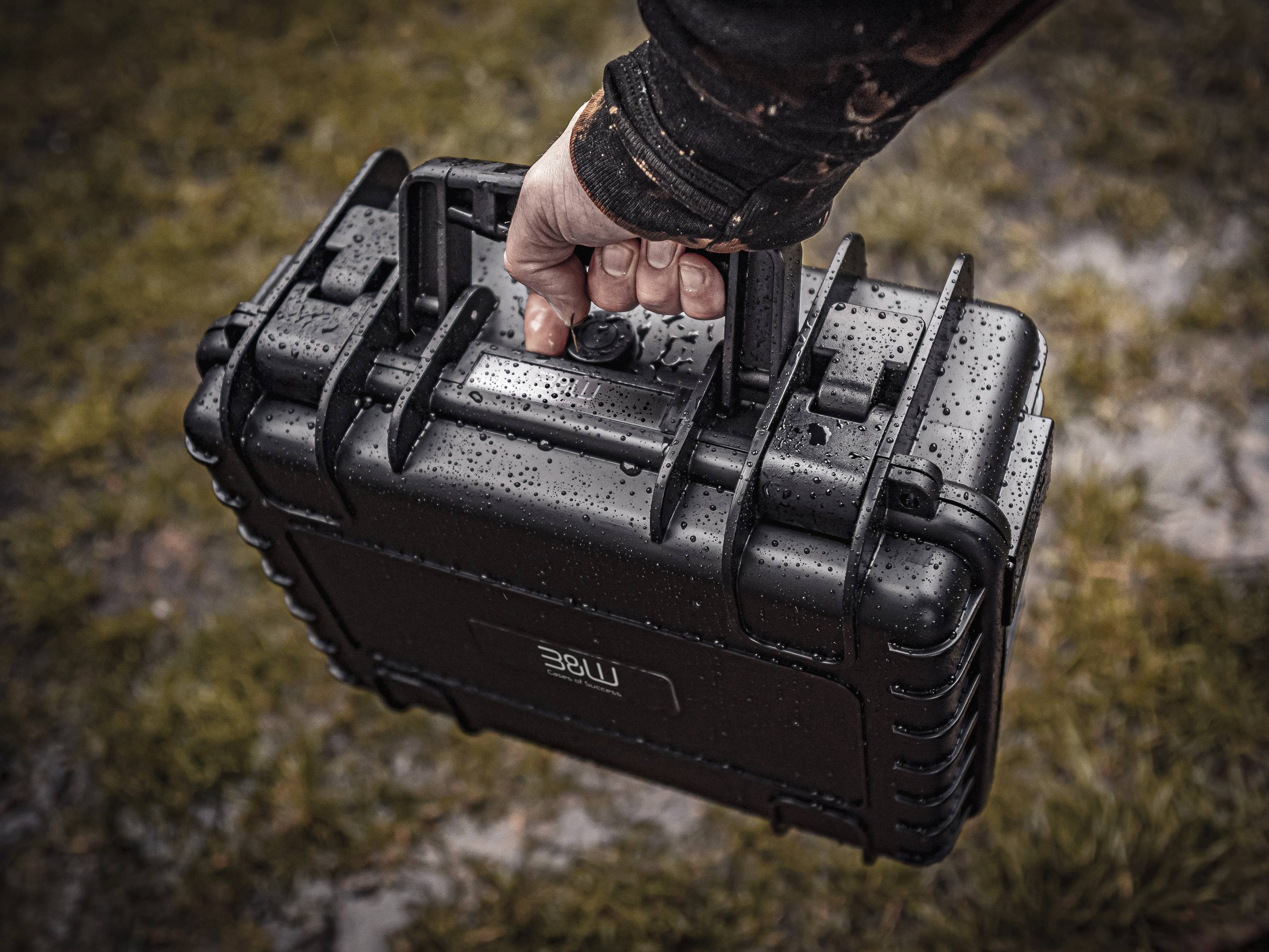 Une main tient un étui noir imperméable par une journée pluvieuse, avec de l'herbe visible en arrière-plan, suggérant une utilisation ou un transport en extérieur.