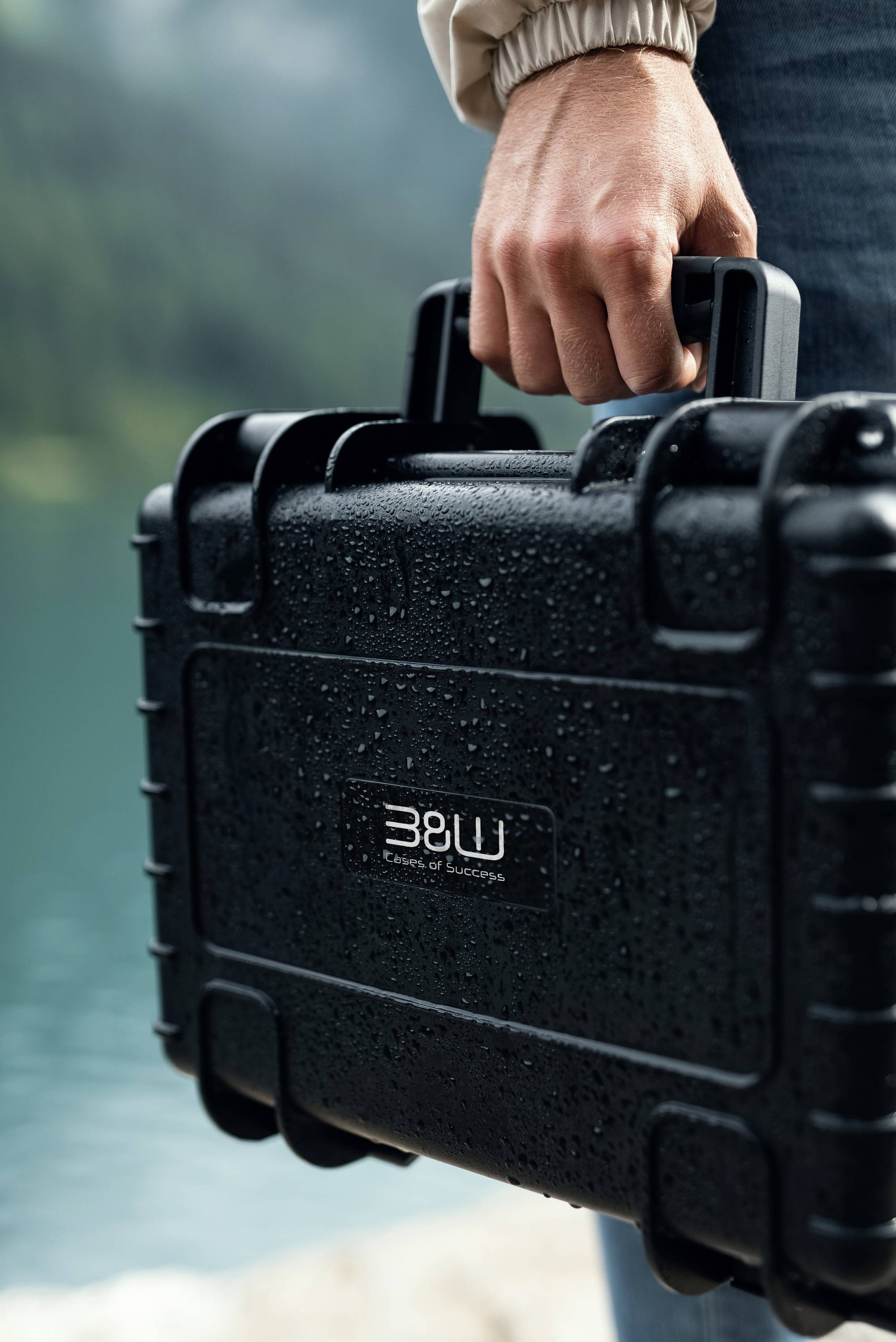 Une personne tenant un étui rigide noir et imperméable près d'un lac, avec le logo "B&W" visible. L'étui est recouvert de gouttes de pluie, suggérant des conditions humides.