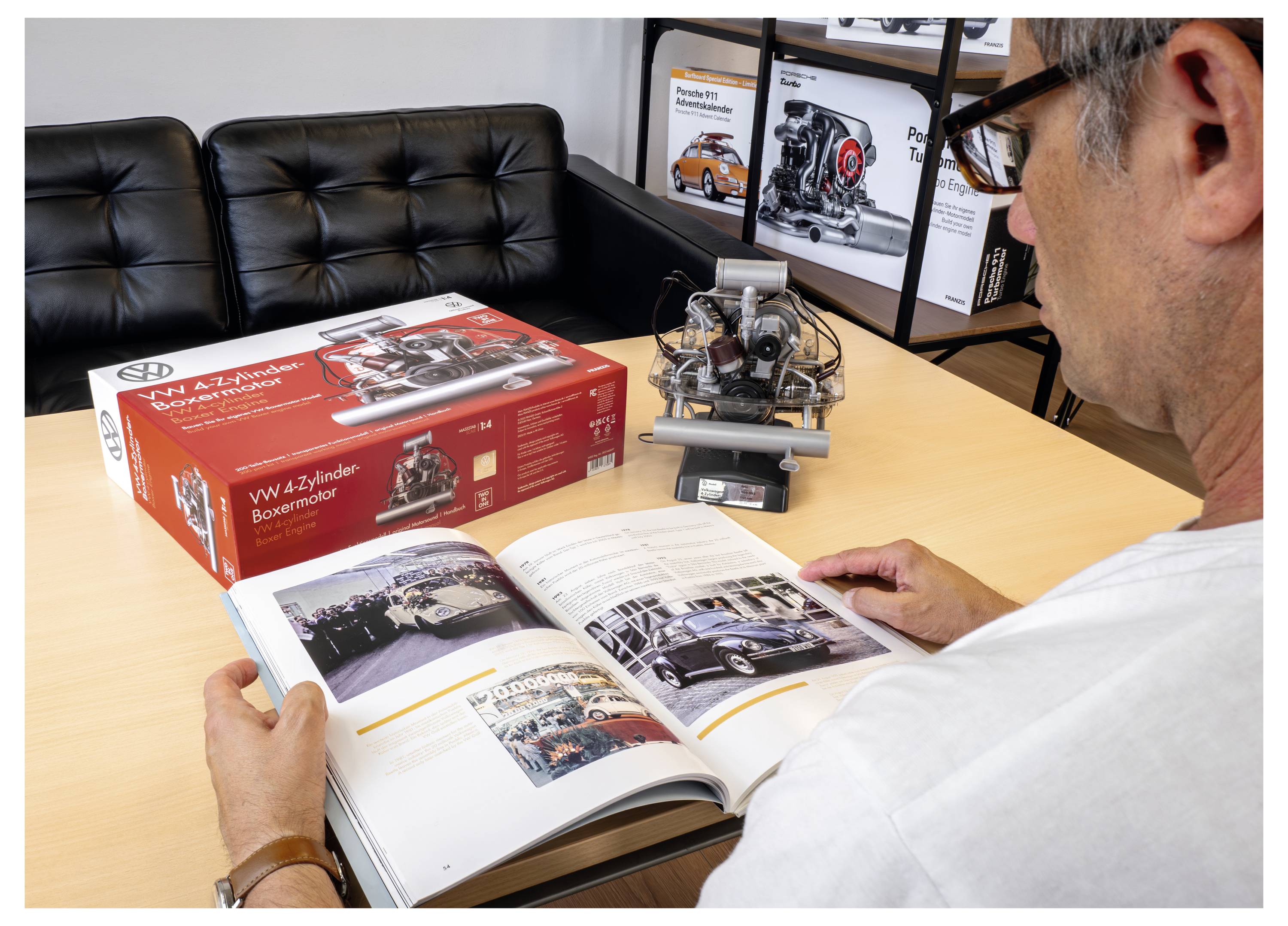 Une personne lit un livre sur l'histoire automobile à une table avec un moteur 4 cylindres VW miniature et sa boîte ; d'autres livres sont visibles en arrière-plan.