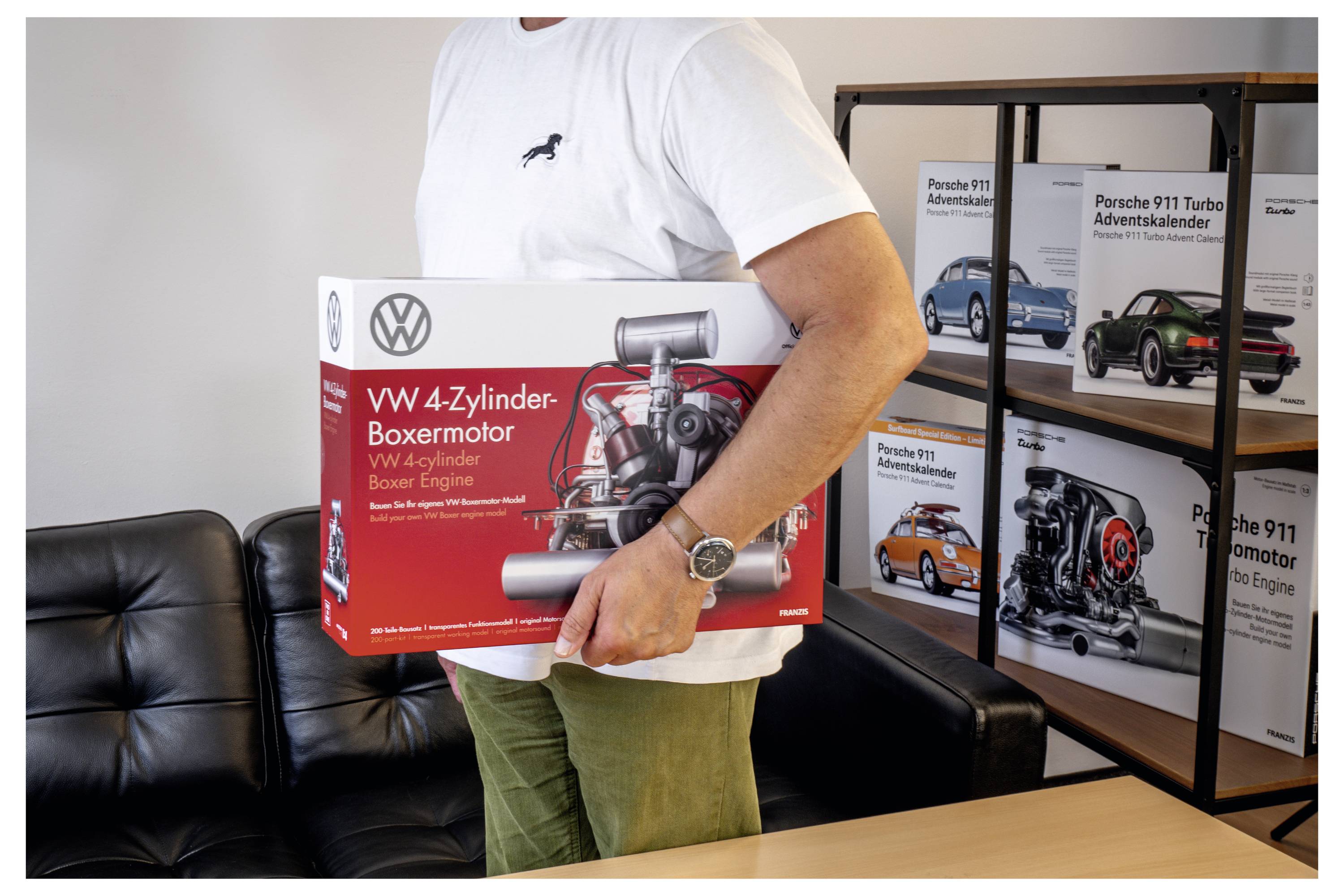 Une personne tenant une boîte rouge étiquetée « VW 4-Zylinder-Boxermotor » dans une pièce décorée avec des boîtes de moteurs miniatures Porsche sur des étagères.
