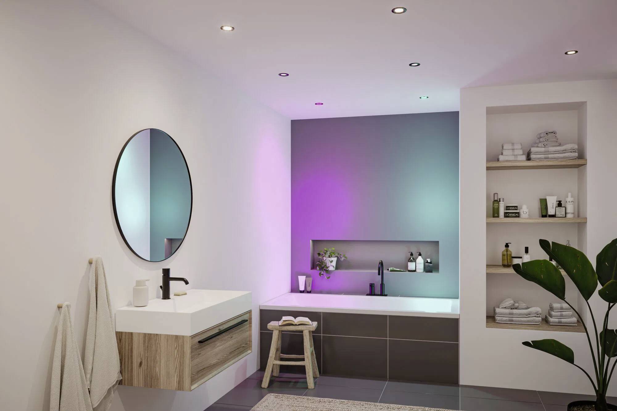 Une salle de bain moderne avec un miroir rond, un lavabo mural, une baignoire sous des lumières colorées, et une étagère avec des serviettes et des produits de toilette.