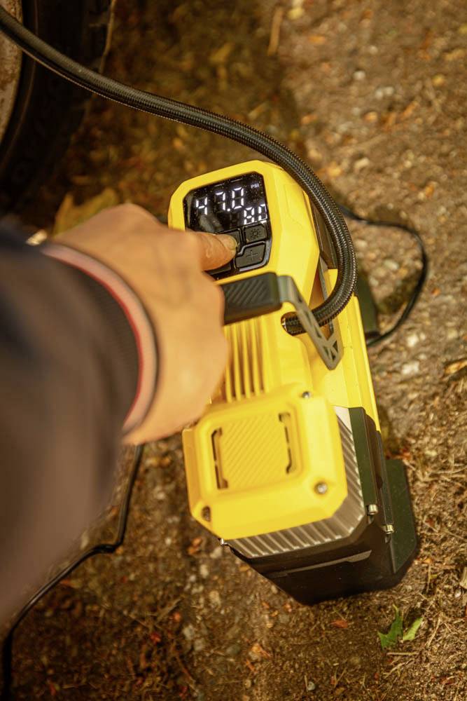 Une personne réglant l'affichage numérique d'un compresseur d'air portable, montrant '4.70', posé sur un sol terreux à côté d'un pneu de véhicule.