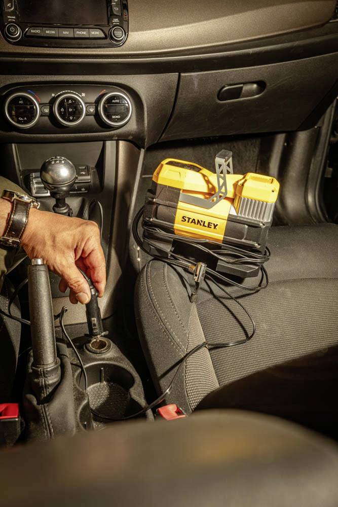 Une personne utilisant un démarreur de batterie à l'intérieur d'une voiture. L'appareil, portant l'étiquette « Stanley », est placé sur le siège passager, et une main le branche sur la prise électrique du véhicule près du levier de vitesses.