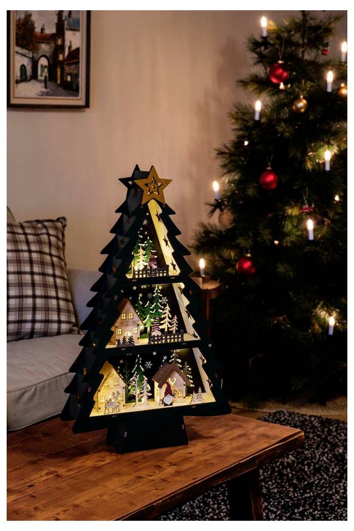 Une décoration de sapin de Noël en bois avec des découpes illuminées de maisons et d'arbres enneigés est posée sur une table près d'un sapin de Noël allumé.