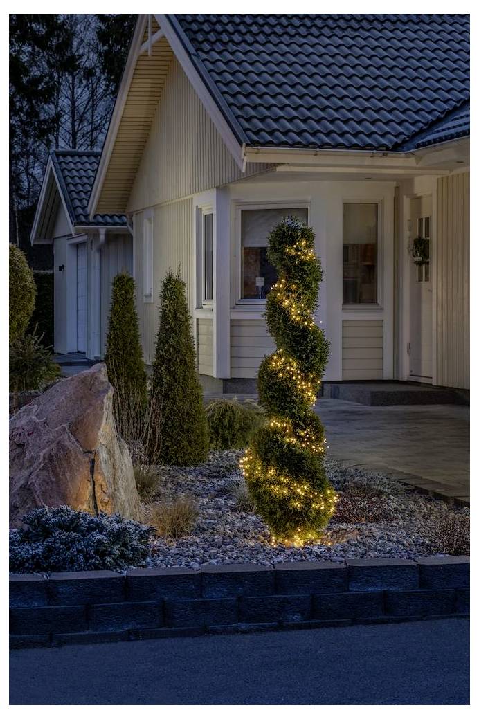 'Un arbre topiaire en spirale avec des lumières scintillantes se dresse illuminé dans la nuit devant une maison, ajoutant un charme décoratif au jardin.'