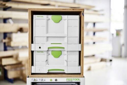 Boîtes de rangement empilées Festool avec poignées vertes dans un atelier. Des étagères avec des planches en bois sont floues en arrière-plan.