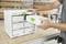 Une personne ouvre un tiroir dans un meuble de rangement Festool blanc, posé sur un établi, entouré d'outils de menuiserie et d'étagères à bois.