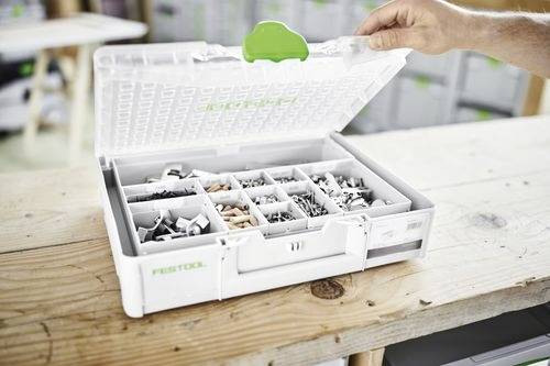 Une boîte à outils blanche ouverte sur une table en bois contient des vis assorties et de la quincaillerie de petite taille, avec la main d'une personne maintenant le couvercle ouvert.
