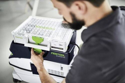 Une personne ajuste un récipient en plastique sur le dessus d'un aspirateur Festool. Le récipient possède une poignée verte et un couvercle transparent.