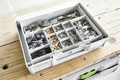 Une boîte à outils ouverte sur une table en bois, remplie de vis assorties, de charnières et de petits goujons en bois rangés dans des compartiments.