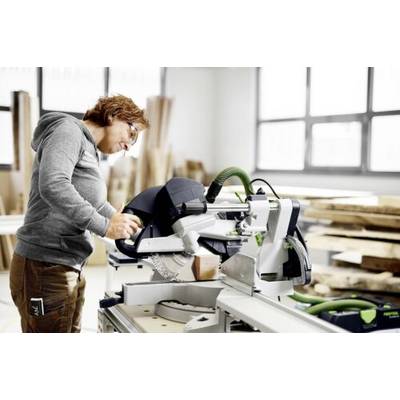 Personne utilisant une scie circulaire sur du bois dans un atelier lumineux, concentrée sur une coupe précise. Des pièces de bois et de grandes fenêtres sont visibles.