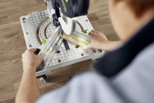 Une personne utilise une scie à onglets sur un sol en bois, en coupant un morceau de bois étiqueté « FESTOOL ». La scie est positionnée sur une base quadrillée.