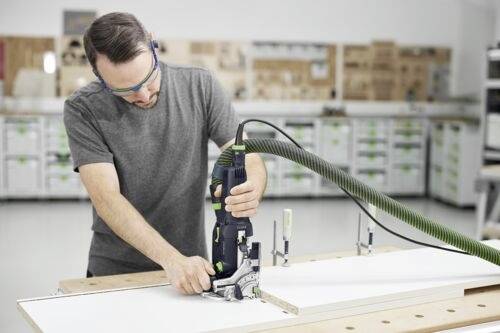Un homme portant des lunettes de sécurité utilise un outil électrique pour couper un morceau de bois dans un atelier ; des étagères de rangement sont visibles en arrière-plan.