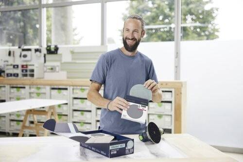 Une personne dans un atelier sourit tout en montrant un paquet de disques de ponçage à côté d'une boîte ouverte, avec des outils visibles en arrière-plan.