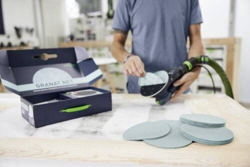 « Une personne change le tampon de ponçage sur une ponceuse électrique, avec une boîte de disques abrasifs Granat Net visible sur un établi dans un atelier. »