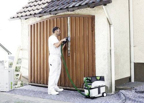 Une personne en combinaison blanche utilise un pistolet de peinture sur une porte de garage en bois. Une machine de peinture avec un tuyau vert est posée au sol à proximité.
