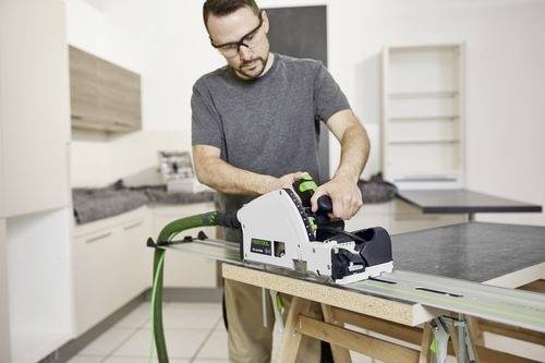 Un homme dans un atelier utilisant une scie électrique sur une planche de bois, entouré d'outils d'atelier et d'étagères, concentré sur une coupe précise.