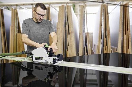 Une personne portant des lunettes de sécurité utilise une scie circulaire pour couper une grande feuille de matériau sombre, avec des piles de panneaux de bois en arrière-plan.