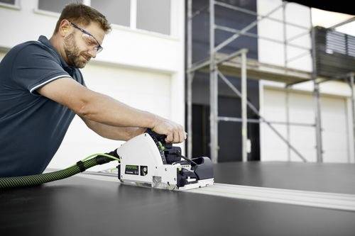 Une personne portant des lunettes de sécurité utilise un outil électrique pour couper un grand panneau noir. Des échafaudages et un bâtiment sont visibles en arrière-plan.