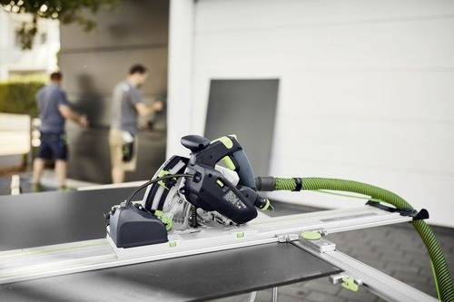Une scie électrique est positionnée sur des rails pour une coupe précise. Deux personnes au second plan flou manipulent de grands panneaux, suggérant un environnement de construction ou d'atelier.