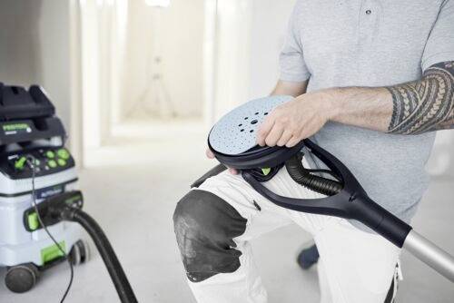 Une personne s'agenouille en changeant la patine d'un outil de ponçage, avec un aspirateur à proximité dans une pièce bien éclairée en cours de rénovation.