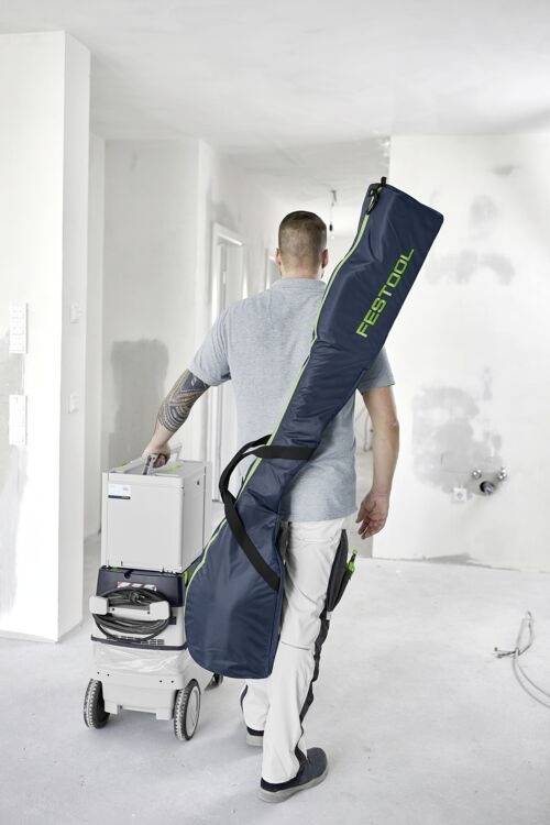 Une personne transportant un grand sac Festool et poussant de l'équipement sur un chariot traverse une pièce partiellement rénovée aux murs blancs.