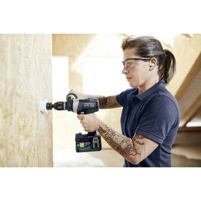 Une personne portant des lunettes de sécurité et une protection auditive utilise une perceuse électrique sur un mur en bois à l'intérieur d'un bâtiment en construction.