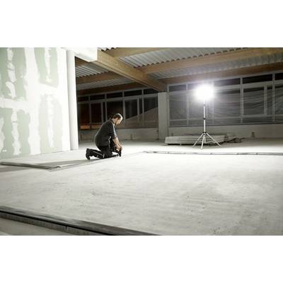 Une personne s'agenouille sur le sol dans une grande pièce partiellement construite, avec des poutres métalliques et un projecteur de chantier lumineux.