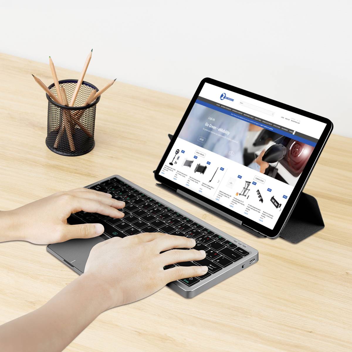 Une personne tape sur un clavier sans fil connecté à une tablette affichant le site web d'un magasin d'électronique en ligne. Un porte-stylos se trouve à proximité.
