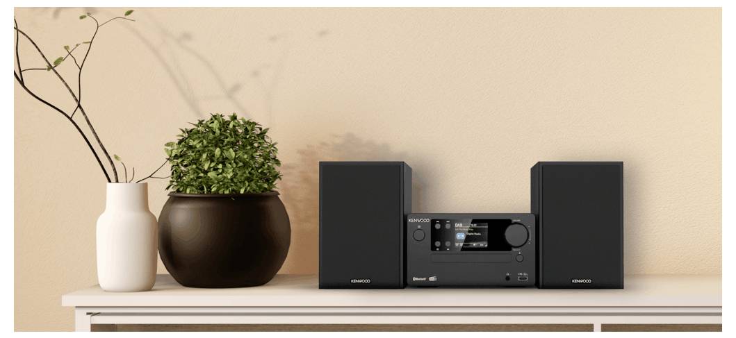 Un système stéréo avec des enceintes est posé sur une étagère à côté de deux vases décoratifs, dont un contenant une plante verte, contre un mur beige.