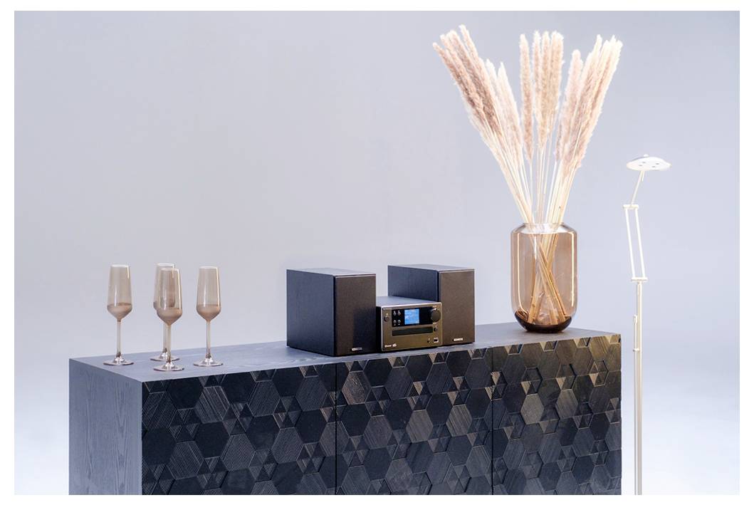Un système stéréo moderne est posé sur un meuble au motif géométrique, à côté de flûtes à champagne et d'un vase rempli d'herbes de la Pampa ; une lampe se tient à proximité.
