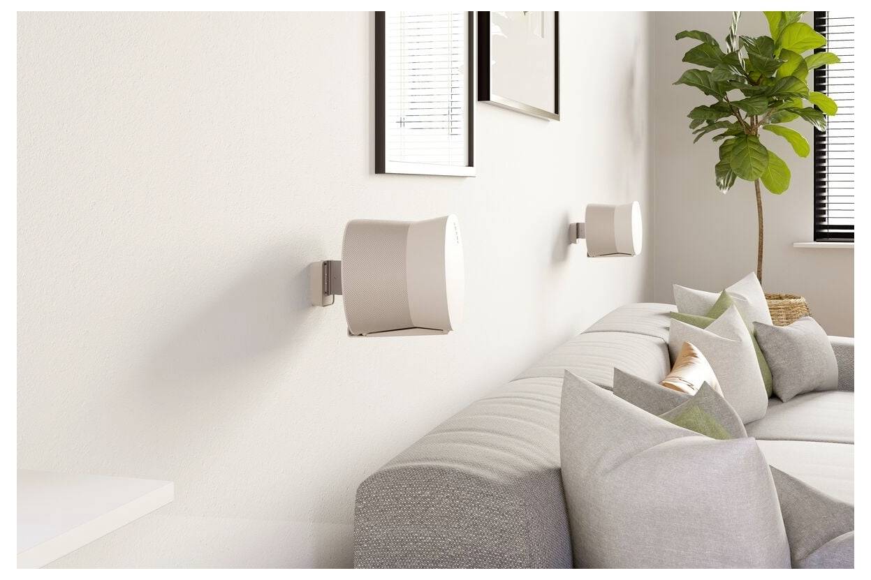 Salon moderne avec un canapé gris clair, des coussins décoratifs, deux enceintes murales, des œuvres encadrées et une plante en pot près de la fenêtre.