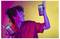 Une personne vêtue d'une chemise rouge sourit en tenant deux tasses colorées, sur un fond vibrant violet, rouge et jaune.