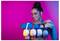 Une femme avec une coiffure en chignon sourit, tenant une rangée d'écouteurs sans fil colorés devant un fond rose et bleu vif.