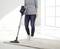 Une personne portant un jean et un pull passe l'aspirateur sur un tapis de couleur claire avec un aspirateur sans fil dans une pièce lumineuse avec de grandes fenêtres.