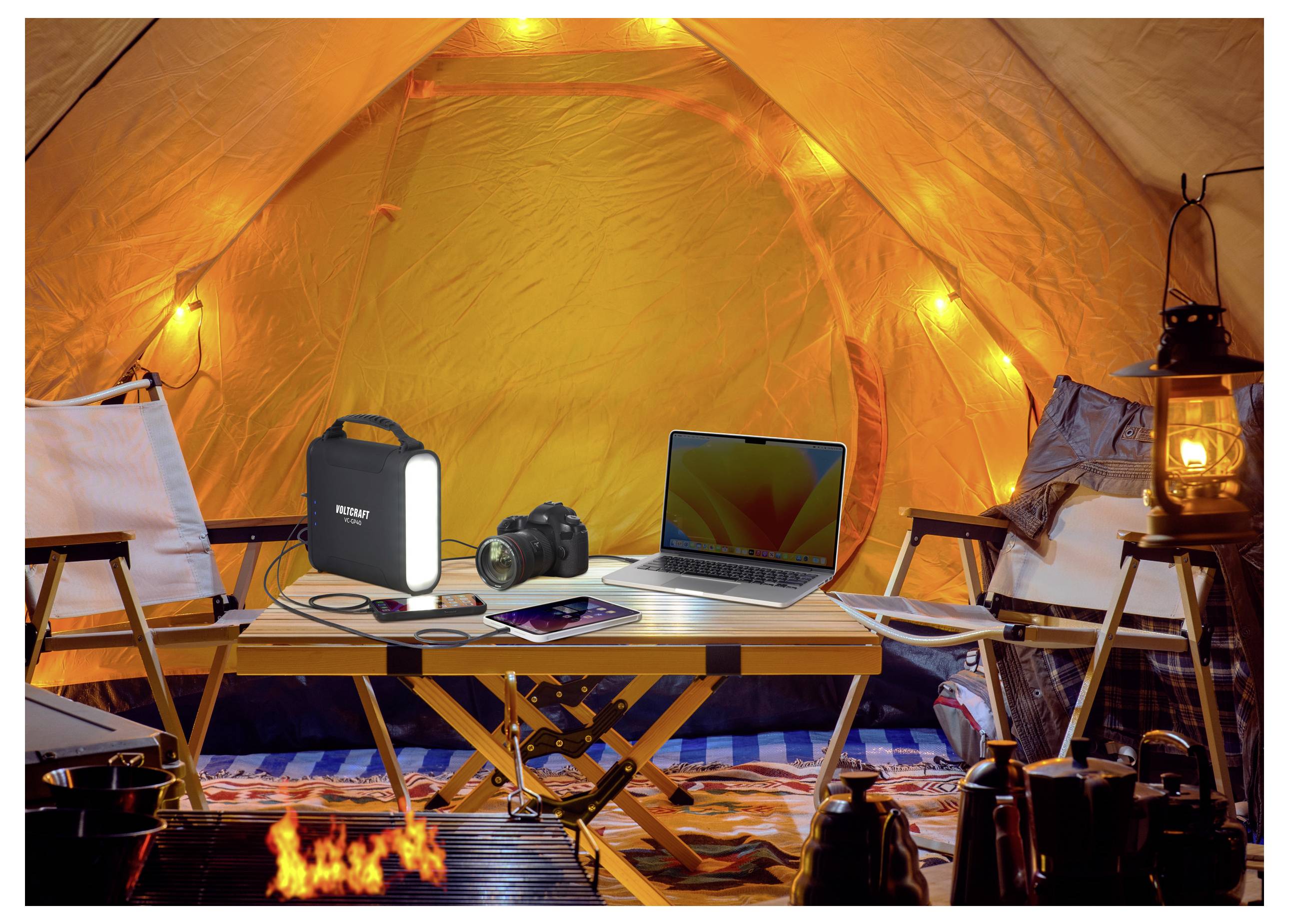 À l'intérieur d'une tente éclairée, une table tient un ordinateur portable, un appareil photo, une batterie externe et une tablette. Des lanternes fournissent une lumière chaleureuse, créant un campement confortable et accueillant.