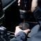 Une personne verse du café d'une tasse de voyage dans une petite tasse à l'intérieur d'une voiture. La tasse est noire avec un bandeau doré près du bord supérieur.