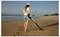 Une personne portant une casquette utilise un détecteur de métaux sur une plage déserte, balayant le sable près de la ligne d'eau alors que le soleil se lève.