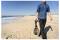 Une personne marche sur une plage sablonneuse en tenant un détecteur de métaux, avec des vagues océaniques à gauche et un ciel bleu clair en arrière-plan.