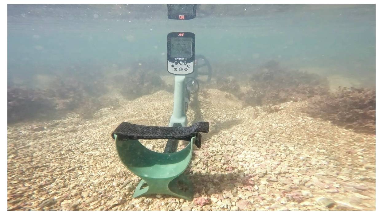 Une vue sous-marine montrant un détecteur de métaux partiellement immergé, balayant un fond marin rocailleux entouré d'algues, indiquant une exploration sous-marine.