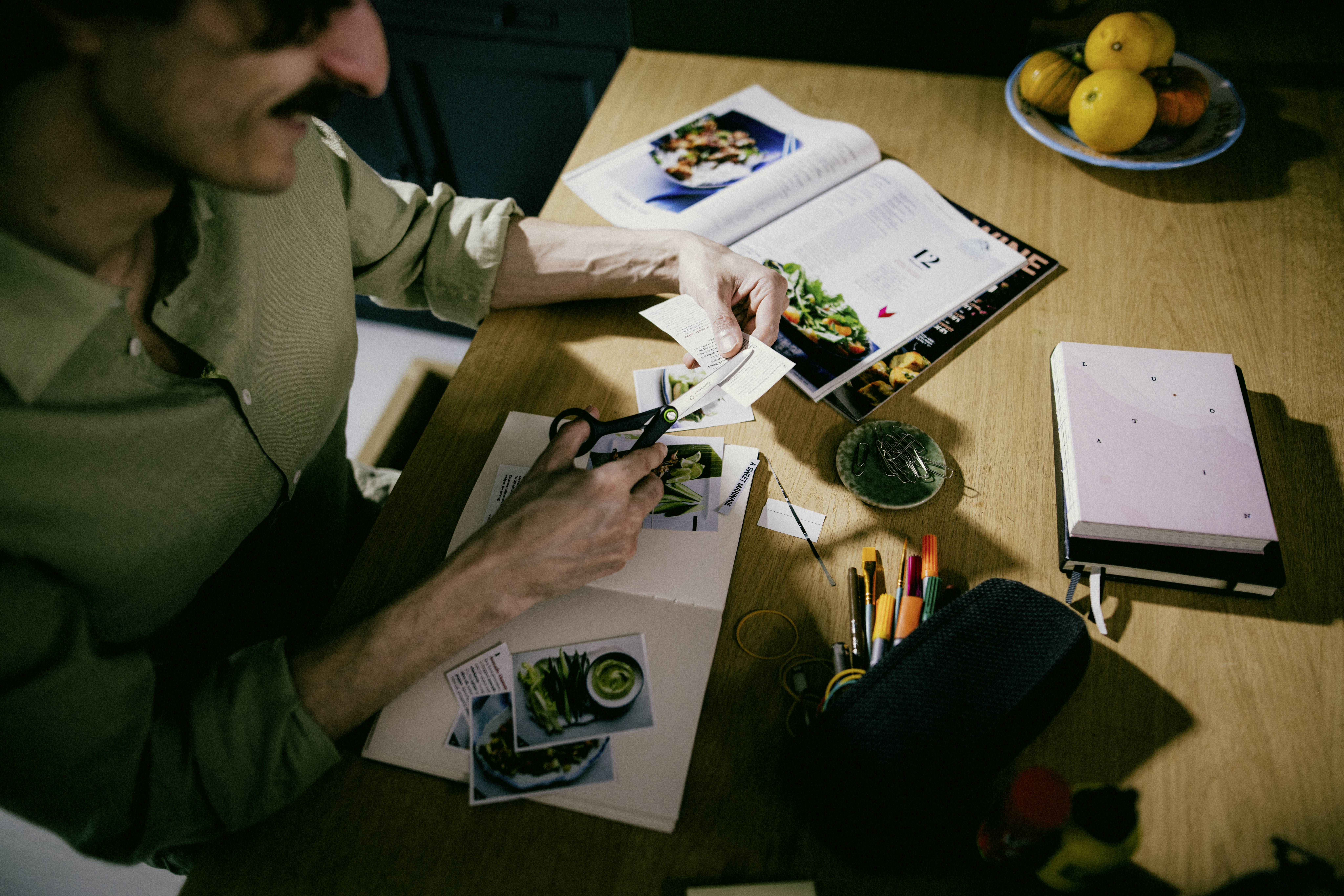 Une personne découpe des photos imprimées à une table avec un livre de cuisine ouvert, des crayons et un bol de fruits, suggérant un projet créatif ou culinaire.