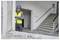 Un ouvrier du bâtiment portant un gilet de sécurité et un casque examine un appareil dans un bâtiment partiellement rénové près d'un escalier.
