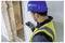 Un ouvrier du bâtiment portant un casque de chantier bleu et un gilet jaune utilise un appareil portable pour mesurer l'épaisseur d'un mur en béton sur un chantier de construction.