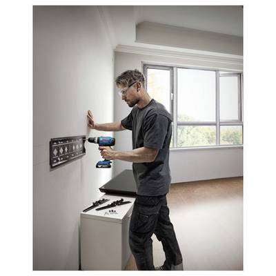 Une personne utilisant une perceuse électrique pour installer un support mural dans une pièce lumineuse avec de grandes fenêtres, portant des lunettes de sécurité et des vêtements de travail décontractés.