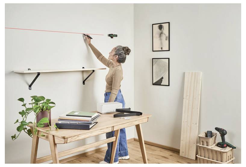 Une personne utilise un niveau laser pour tracer une ligne sur un mur. Une étagère est installée en dessous, et une table avec des livres, une plante et des outils se trouve à proximité.