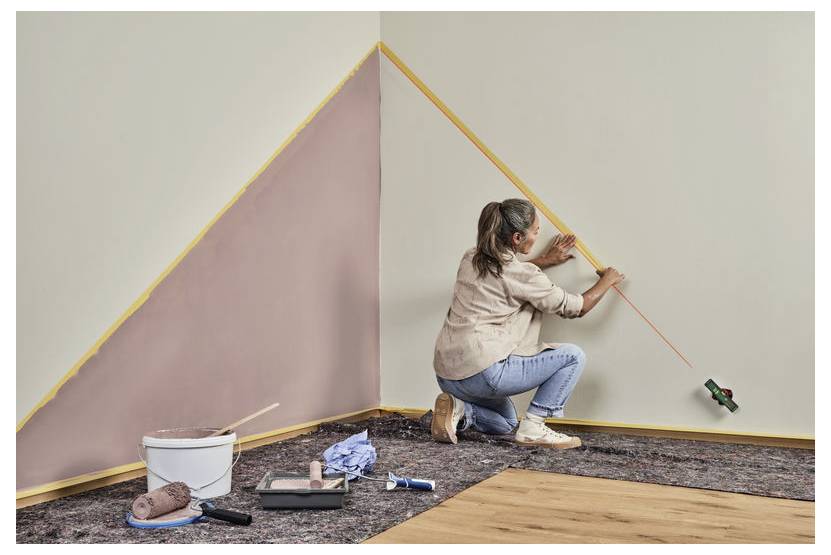 Une femme est agenouillée sur le sol, peignant une section murale géométrique en forme de triangle en rose, à l'aide d'un rouleau et de ruban de masquage.
